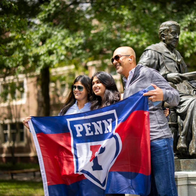 Penn student posing for photo with two family members while holding up flag with the Penn Logo