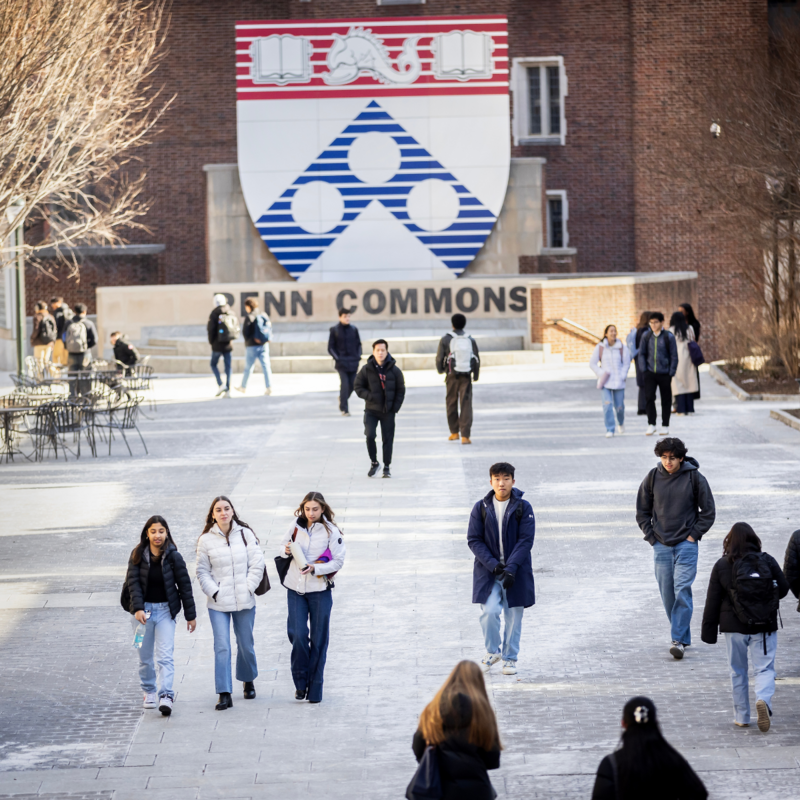 Multiple groups of students wearing large jackets walking through Penn Commons with a large Penn Shield in the background. 