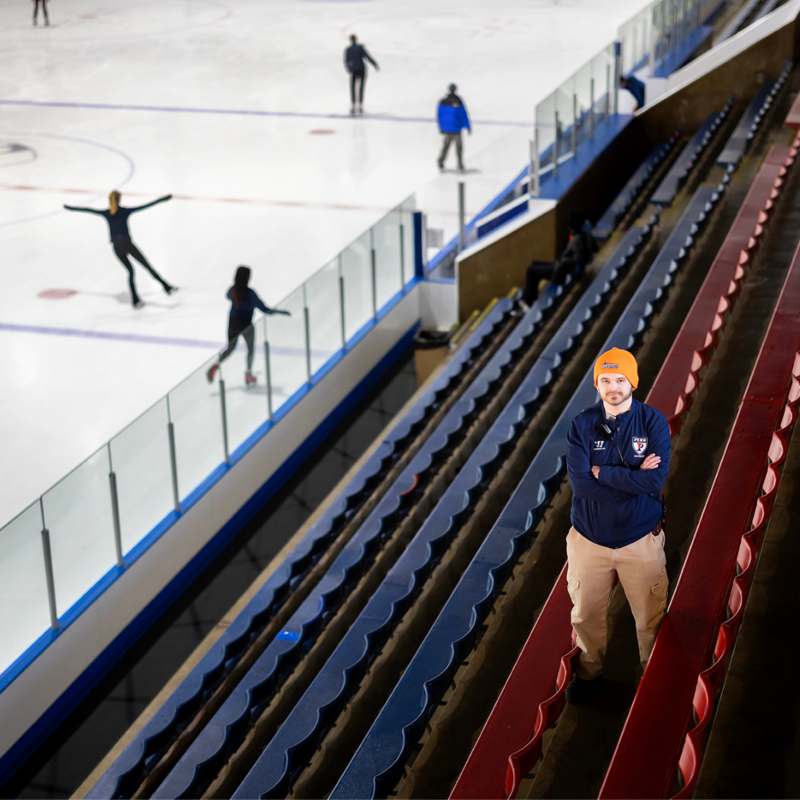 Mike Petrocelli from the Penn Ice Rink standing with his arms folding posing for a photo with multiple people ice skating in the background.