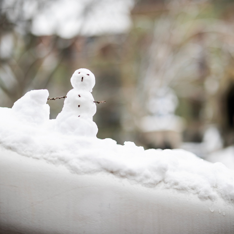 Small snow man on Penn's campus with sticks for arms and pebbles formed to make a face with a blurred college hall in the background.